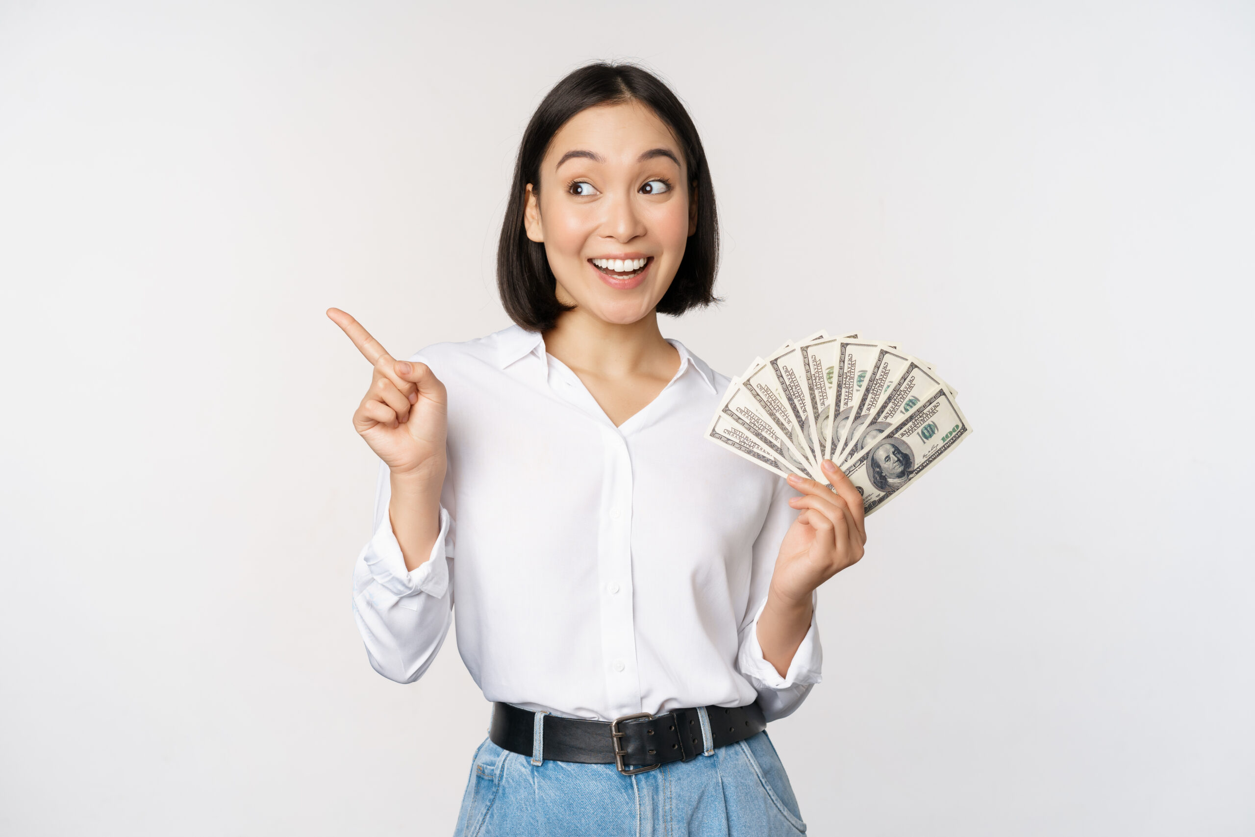 Smiling young modern asian woman, pointing at banner advertisement, holding cash money dollars, standing over white background.